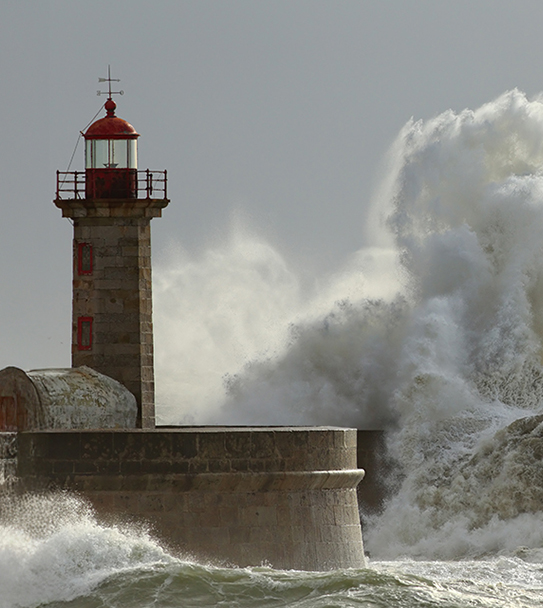A tall, weathered stone lighthouse with a red lantern room stands firmly on a pier as enormous, foamy white waves crash against the rocks and the pier structure during a storm. The sky is overcast and gray, emphasizing the dramatic power of the turbulent sea.