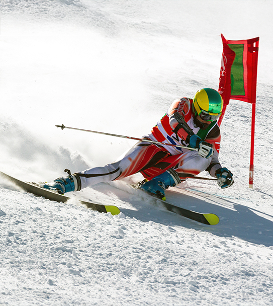 A skier in a red, white, and blue suit, yellow and green helmet, and blue boots carves down a snowy slope past a red and green gate.