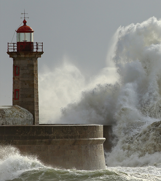 A tall stone lighthouse with a red lantern room stands on a seawall as massive ocean waves crash dramatically against it, sending spray high into the air under a cloudy sky.